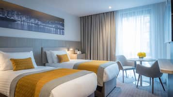 A hotel room with two beds, a desk, a chair, a table with flowers, and a cityscape view.