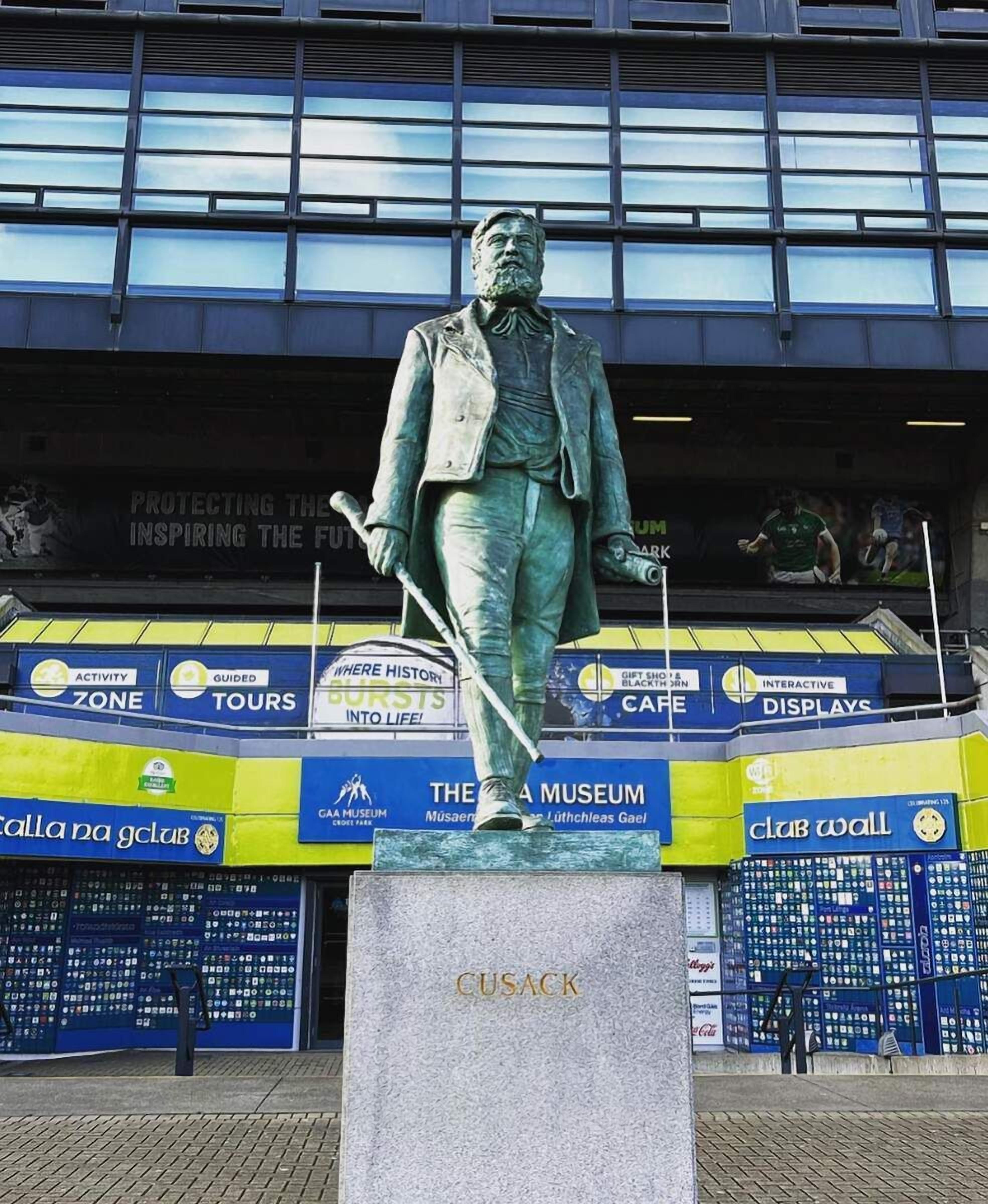 Una statua di un uomo in abito che tiene un battente da baseball, posata su un piedistallo con il nome 'CUSACK' inciso, di fronte a un edificio con diverse pubblicità.
