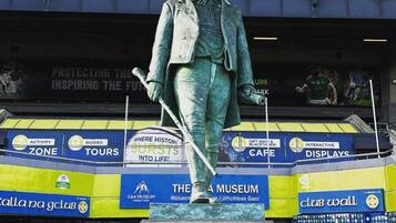 Una statua di un uomo in abito che tiene un battente da baseball, posata su un piedistallo con il nome 'CUSACK' inciso, di fronte a un edificio con diverse pubblicità.