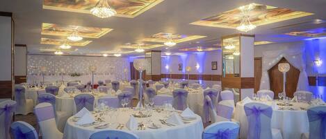 Un salón de banquetes con mesas redondas dispuestas para comer, cada una con mantel blanco y fajín azul.