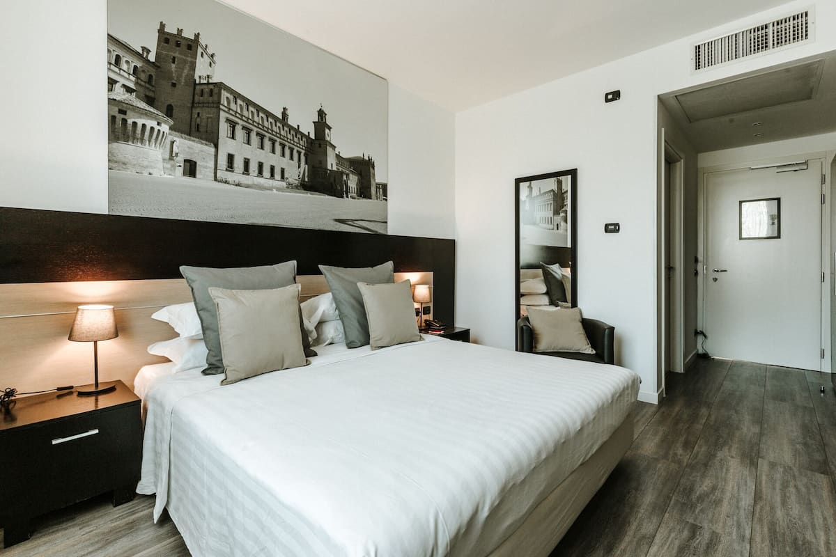Une chambre d’hôtel avec un grand lit, une tête de lit ornée d’une photo représentant un paysage urbain, une table de chevet avec une lampe et un miroir au mur.