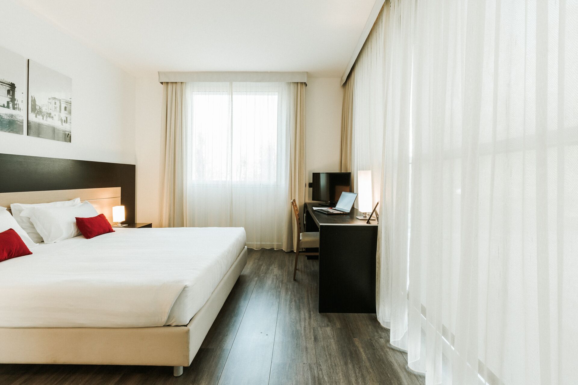 Et hotelværelse med en stor seng, et skrivebord med en bærbar computer, et fjernsyn og et vindue med let gardin.