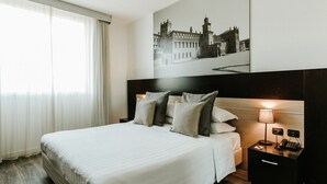 A hotel room with a large bed, a headboard with a light fixture, a nightstand with a lamp, and a framed picture on the wall. - Hotel Carpi (Carpi)