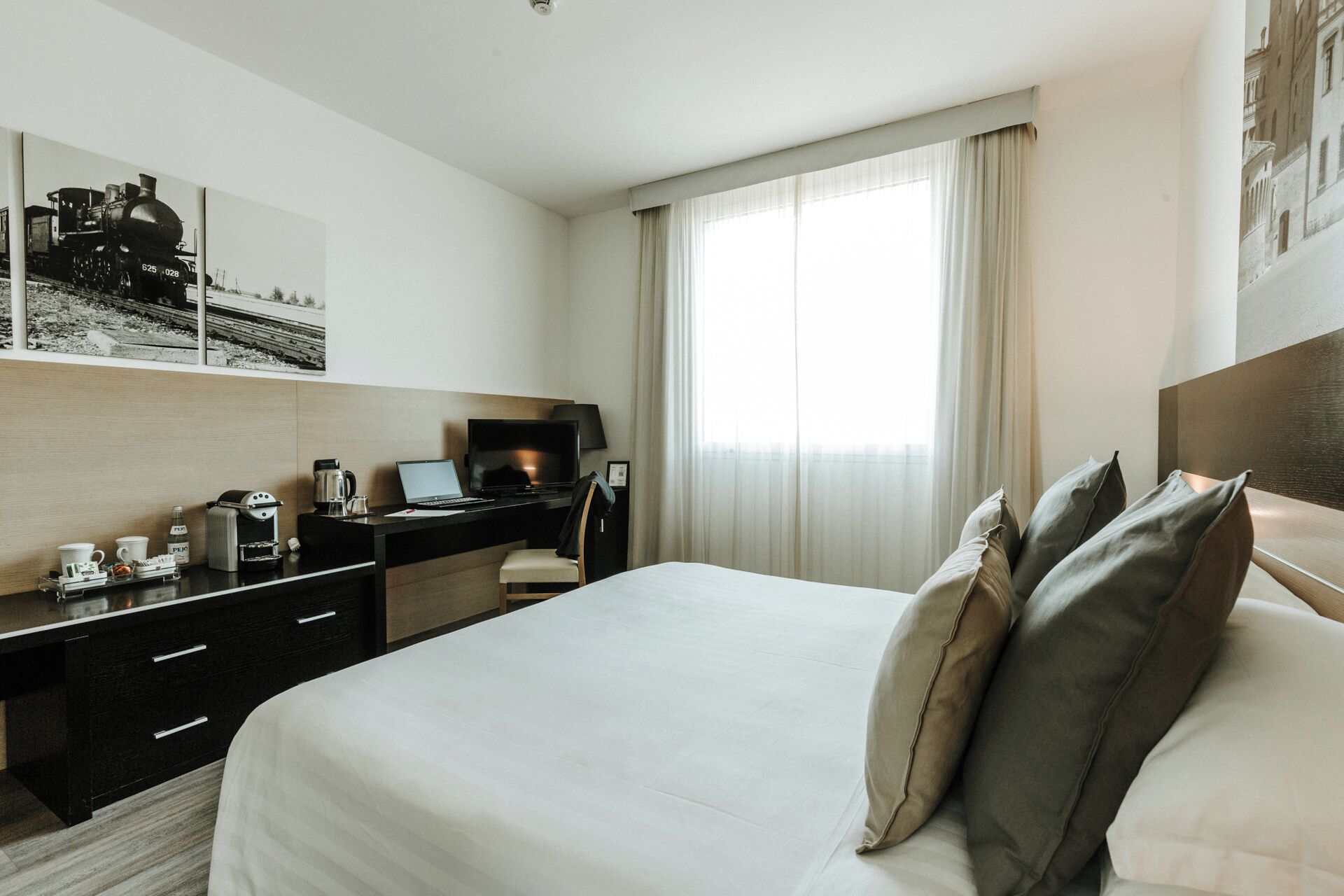 Hotellihuone, jossa on sänky, työpöytä kannettavalla tietokoneella, kahvinkeitin ja seinällä oleva kehystetty kuva.