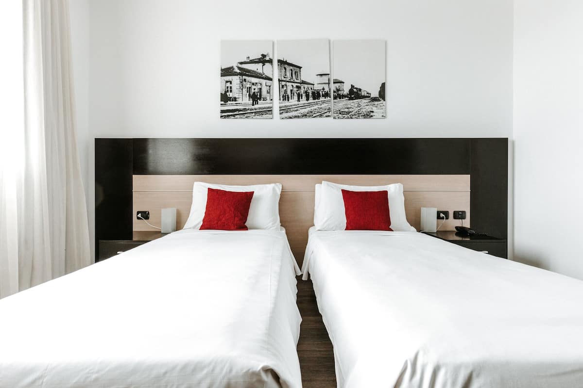 Une chambre d’hôtel avec deux lits, une tête de lit en bois, une table de chevet avec un téléphone et un tableau représentant une gare, accroché au mur.
