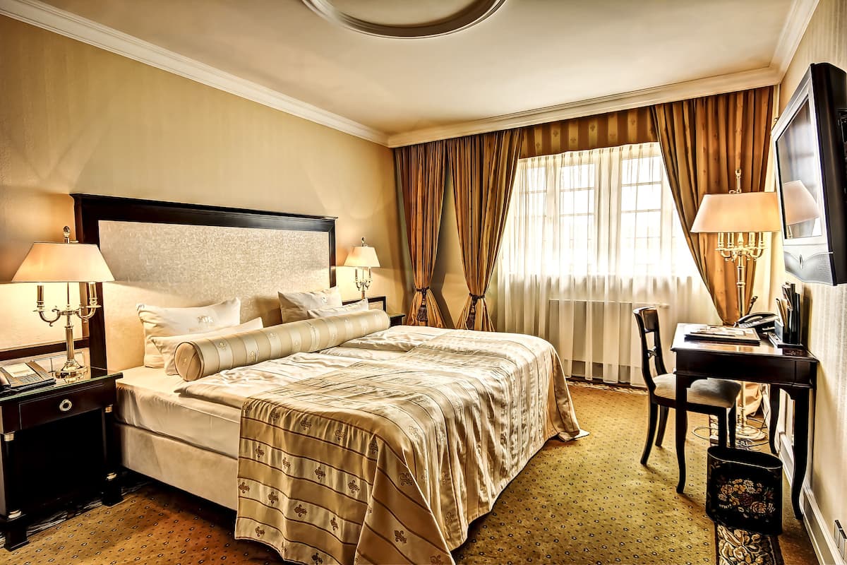 A hotel room with a large bed, two bedside tables with lamps, a desk with a chair, a television, and a window with curtains.