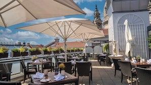 Un área de comedor al aire libre con mesas y sillas, grandes sombrillas y vistas a un edificio histórico con una torre del reloj.