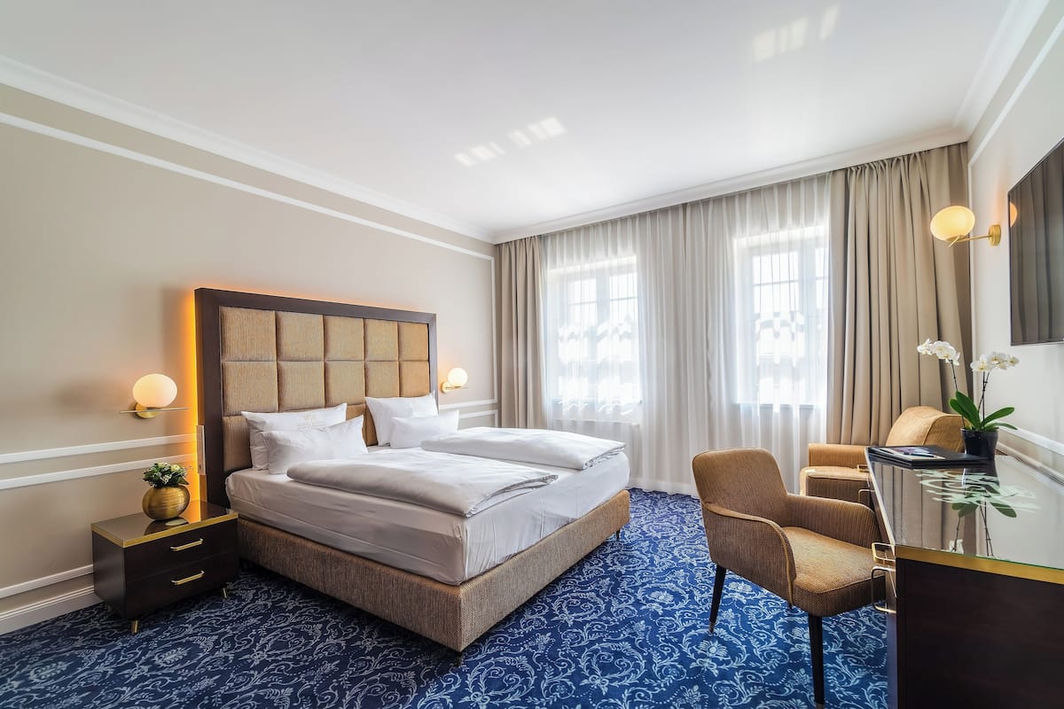 A hotel room with a large bed, a desk, and a chair. There is a nightstand with a lamp and a potted plant.