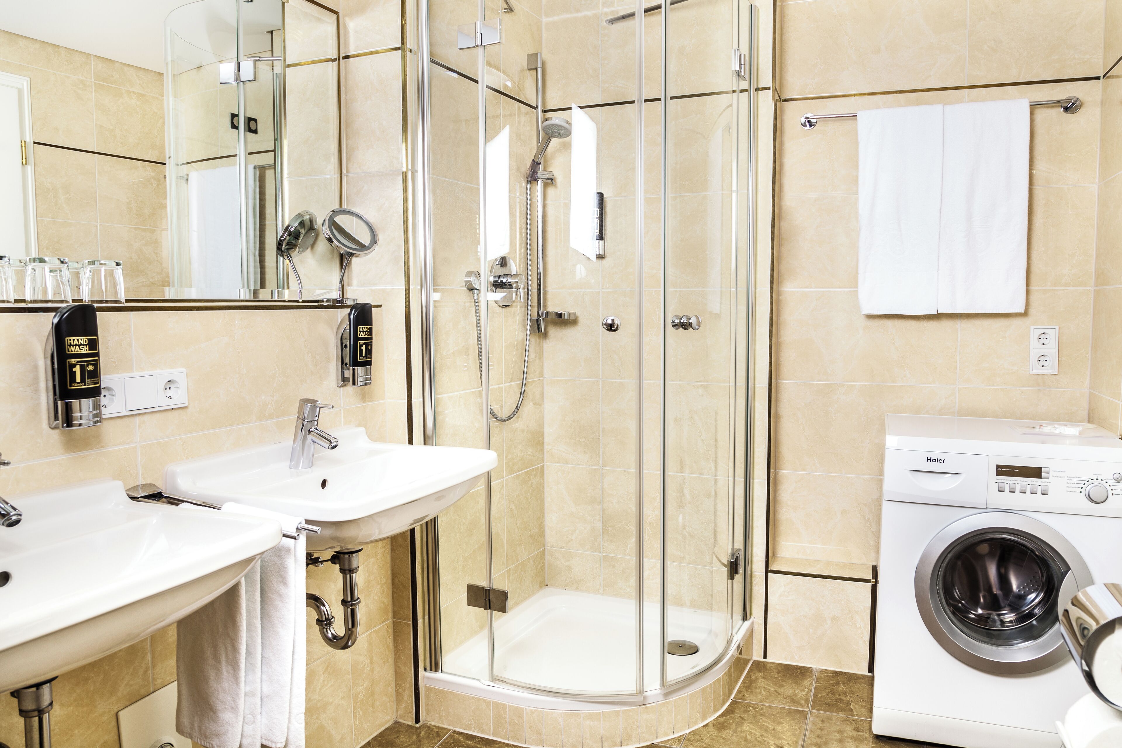 Un baño con dos lavamanos, ducha con mampara de vidrio y lavadora.