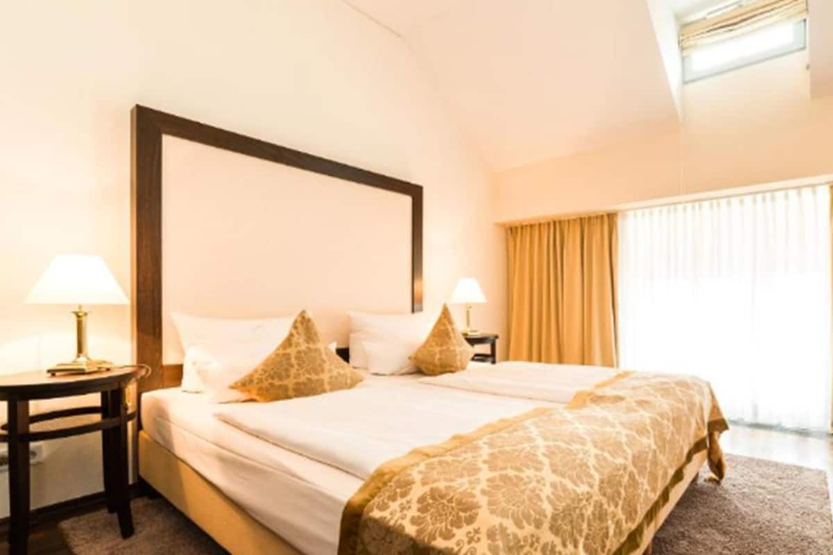 A hotel room with a large bed, two bedside tables with lamps, a window with curtains, and a wooden floor.
