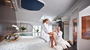 Una pareja vestida de batas blancas sentada en un banco con mosaicos en un moderno espacio de spa, con techo estrellado y piscina cubierta de cristal.