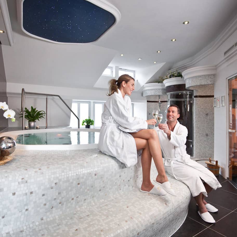 A couple in white robes sitting on a mosaic-tiled bench in a modern spa area with a starry ceiling and a glass-enclosed pool.