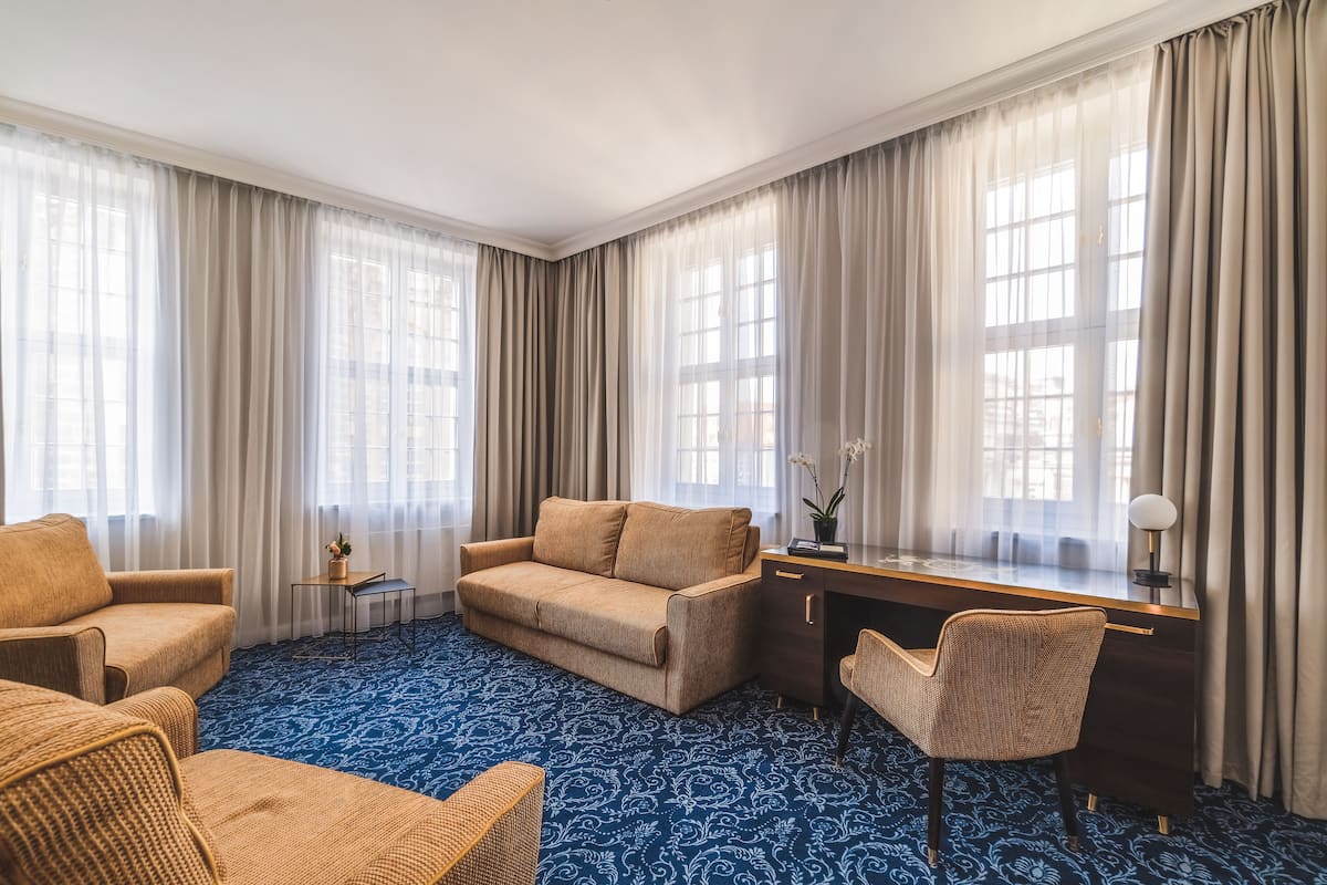A hotel room with a blue patterned carpet, a beige sofa, a wooden desk, and a wicker chair.