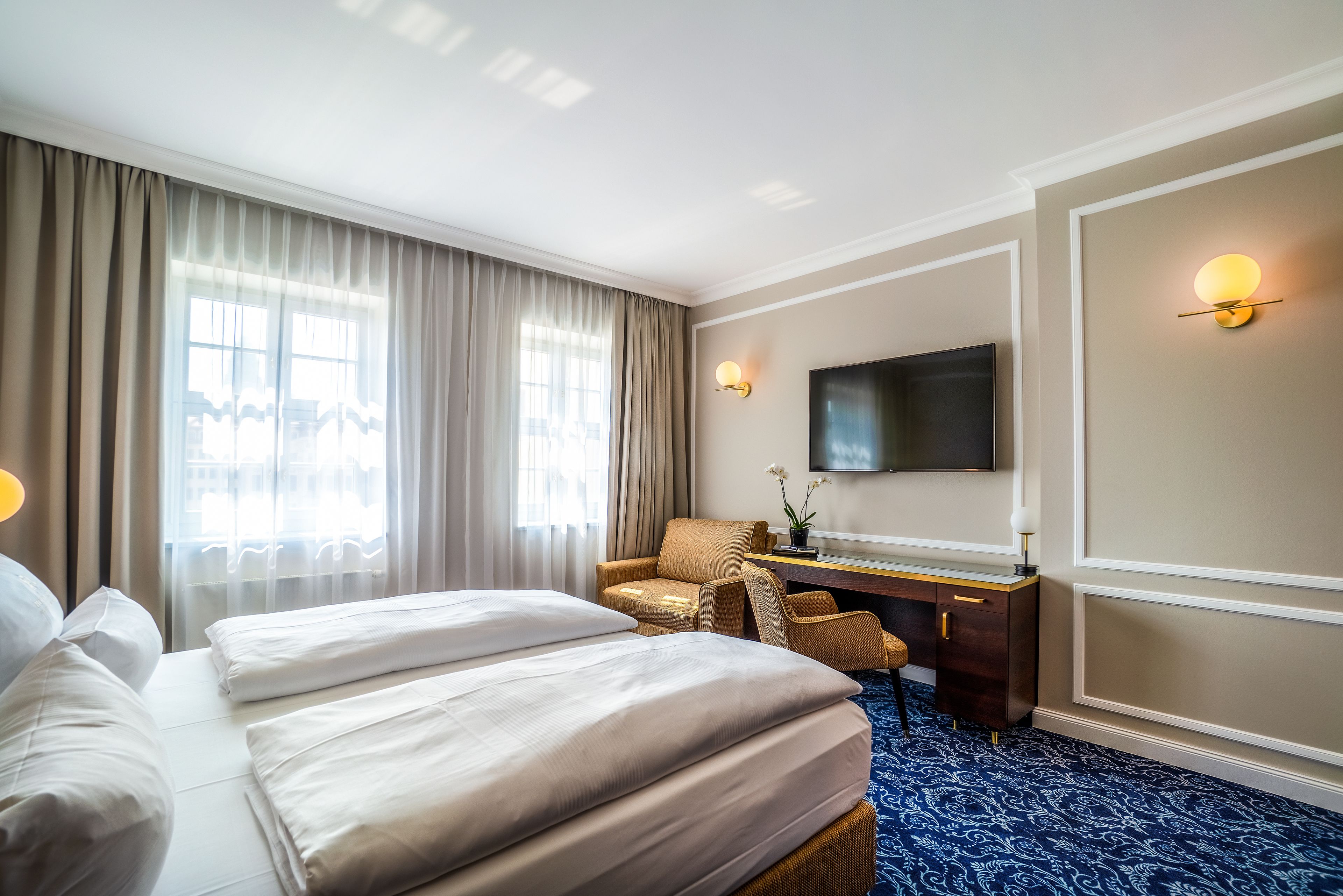 A hotel room with two beds, a desk, a chair, a TV, and a large window with curtains.