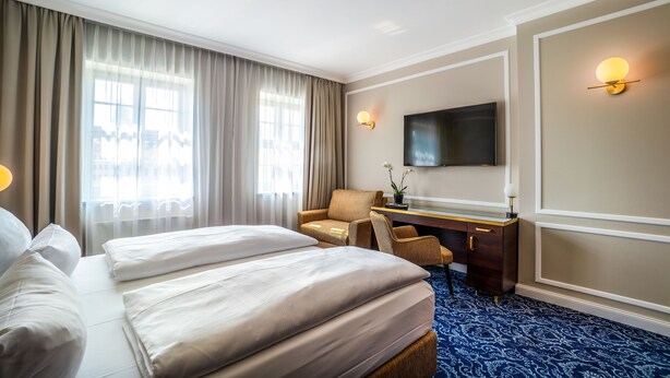 A hotel room with two beds, a desk, a chair, a TV, and a large window with curtains.