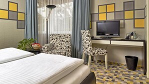 A hotel room with a bed, a desk with a TV, two chairs, a plant, and a trash bin. - Hotel & Restaurant 5 Linden (Südharz)