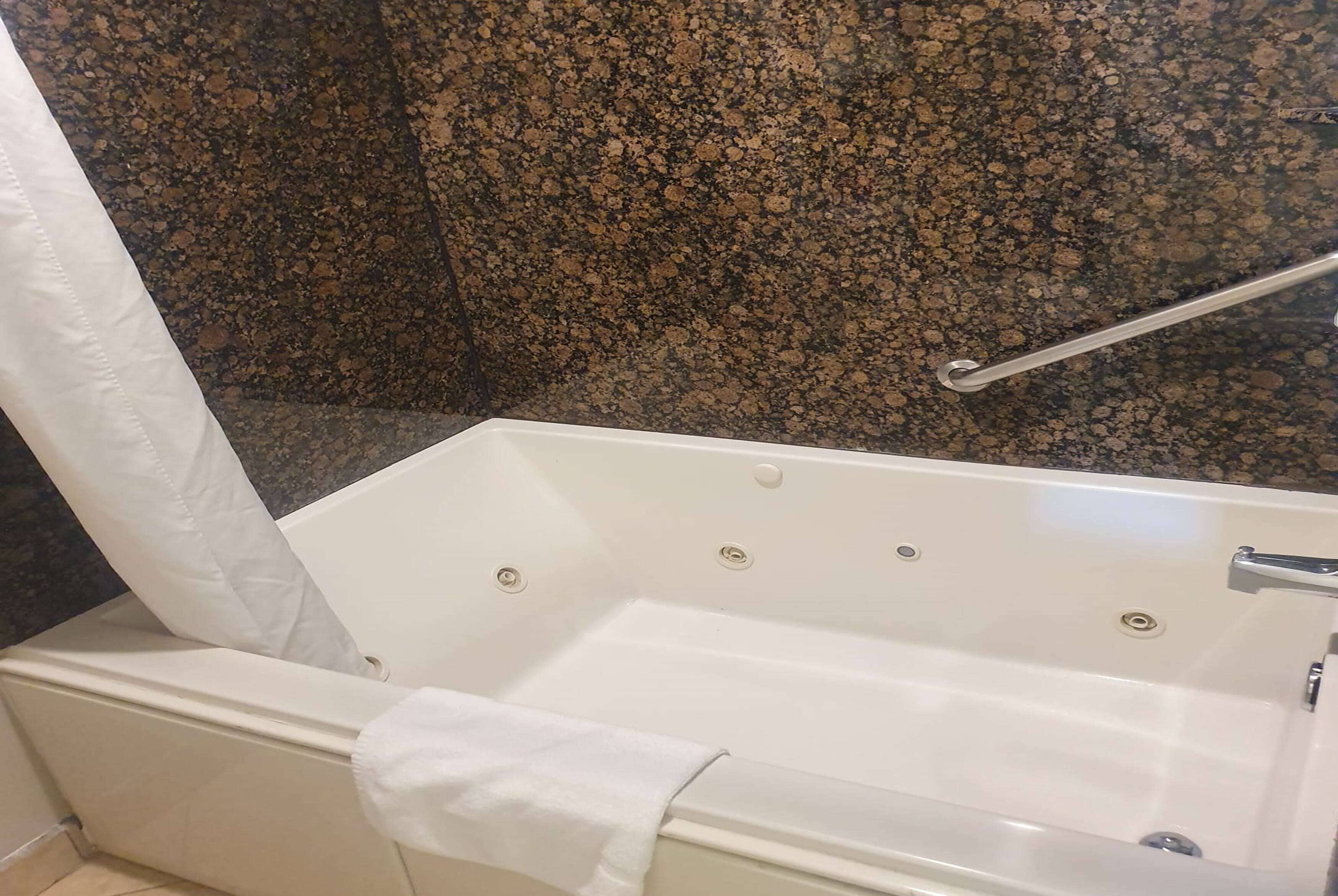 Una bañera con una toalla blanca, una barra de metal para agarrarse y una pared de granito.