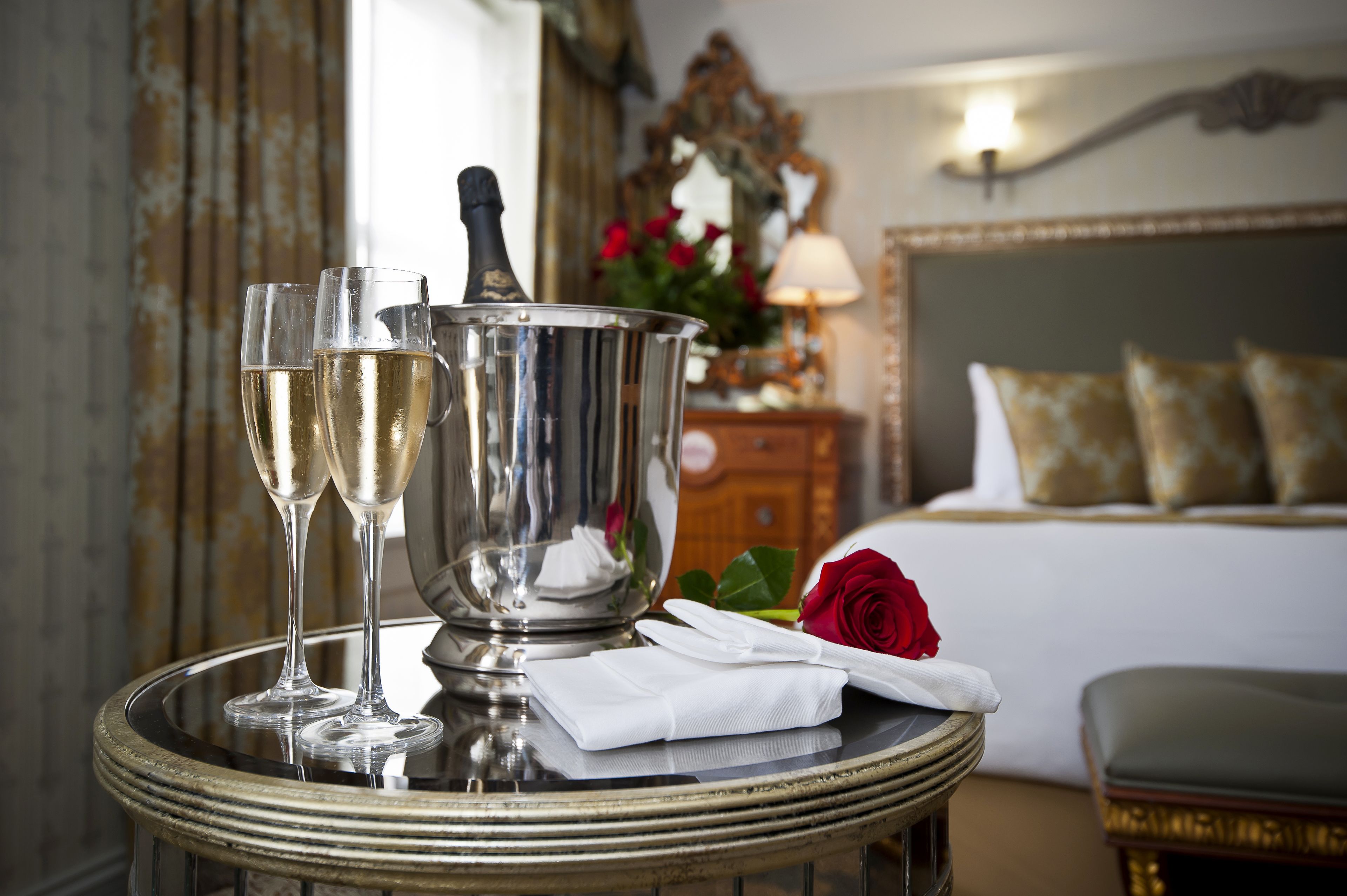 A room with a bed, a bedside table with a bottle and glasses, and a red rose.