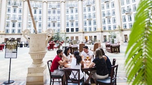 Un'ampia area interna con soffitto alto e grandi finestre, caratterizzata da un colonnato decorativo centrale e una zona pranzo con tavoli e sedie.