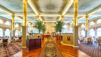 A grand hotel lobby with a central reception desk, ornate columns, and a patterned carpet.