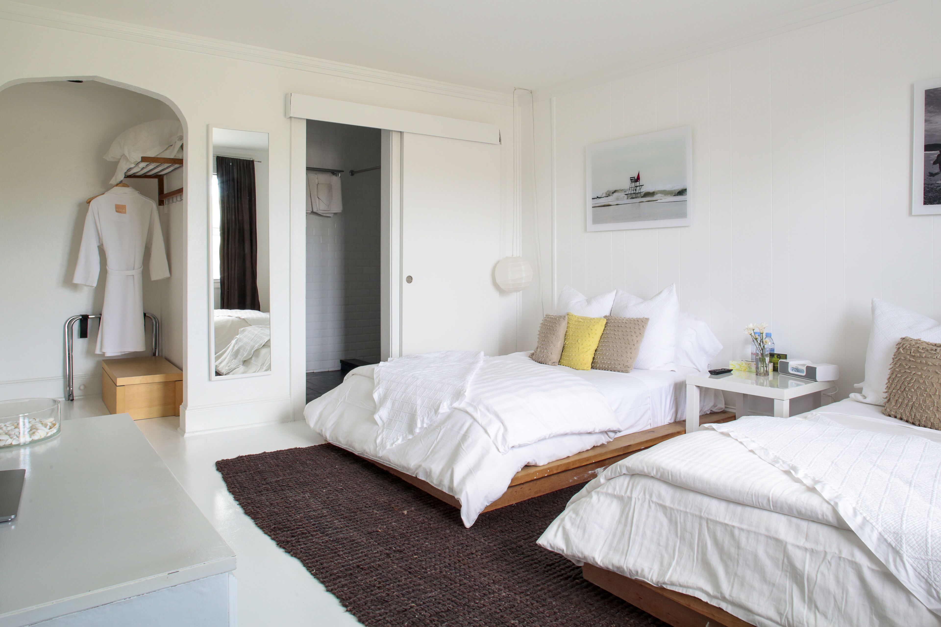 Quarto com duas camas, mesa de cabeceira, espelho, guarda-roupa e banheiro visível por uma porta aberta.