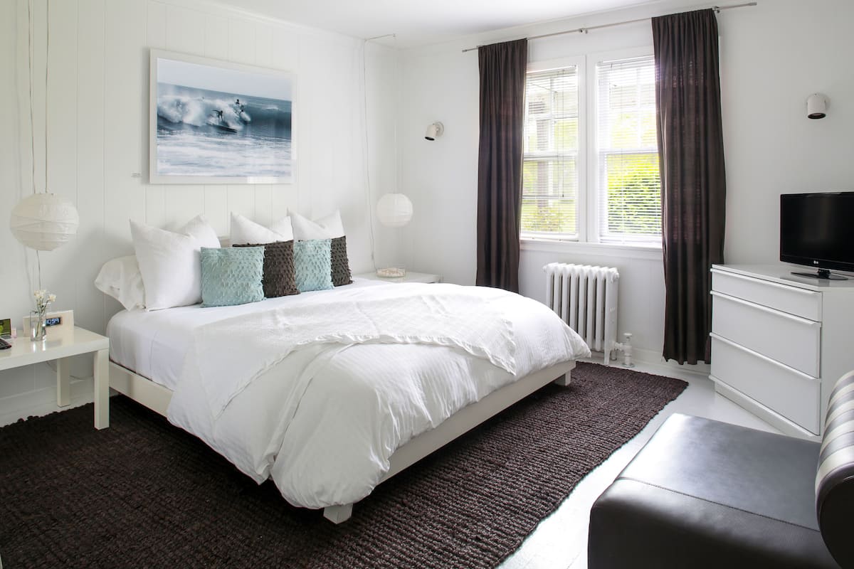 Une chambre à coucher avec un grand lit, une télévision, une table de chevet, une commode, une fenêtre avec des rideaux et un radiateur.