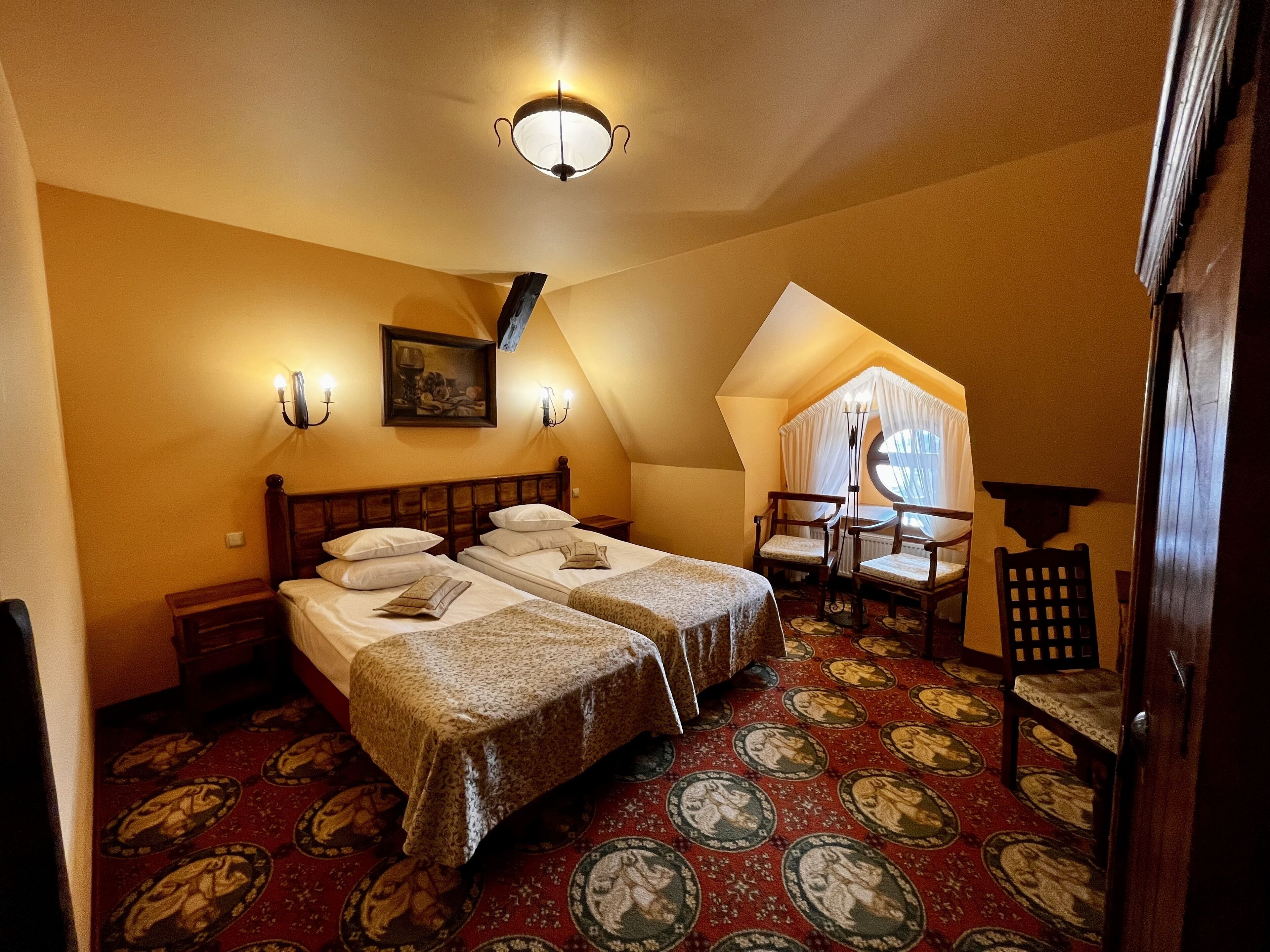 A bedroom with two beds, a small table, and a chair. There is a painting on the wall and a ceiling light fixture.