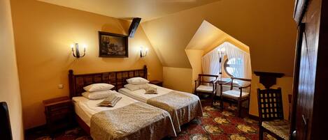 A bedroom with two beds, a small table, and a chair. There is a painting on the wall and a ceiling light fixture.