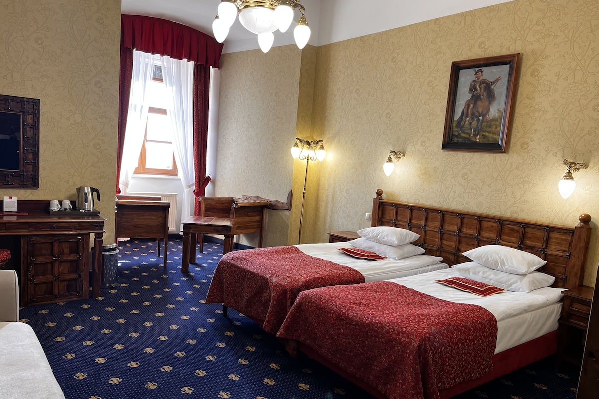 A hotel room with two beds, a desk, a chair, a radiator, and a painting on the wall.