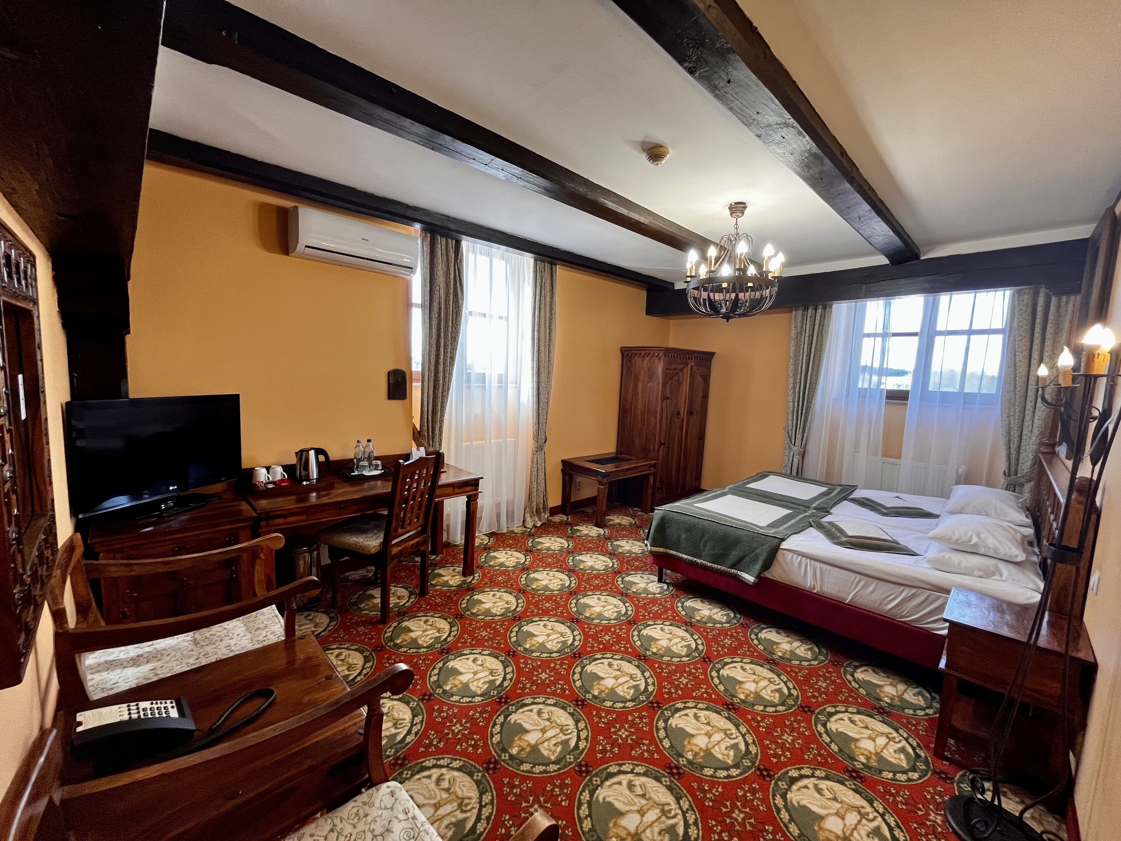 A hotel room with a large bed, a desk with a television, a wardrobe, and a carpet with a traditional pattern.