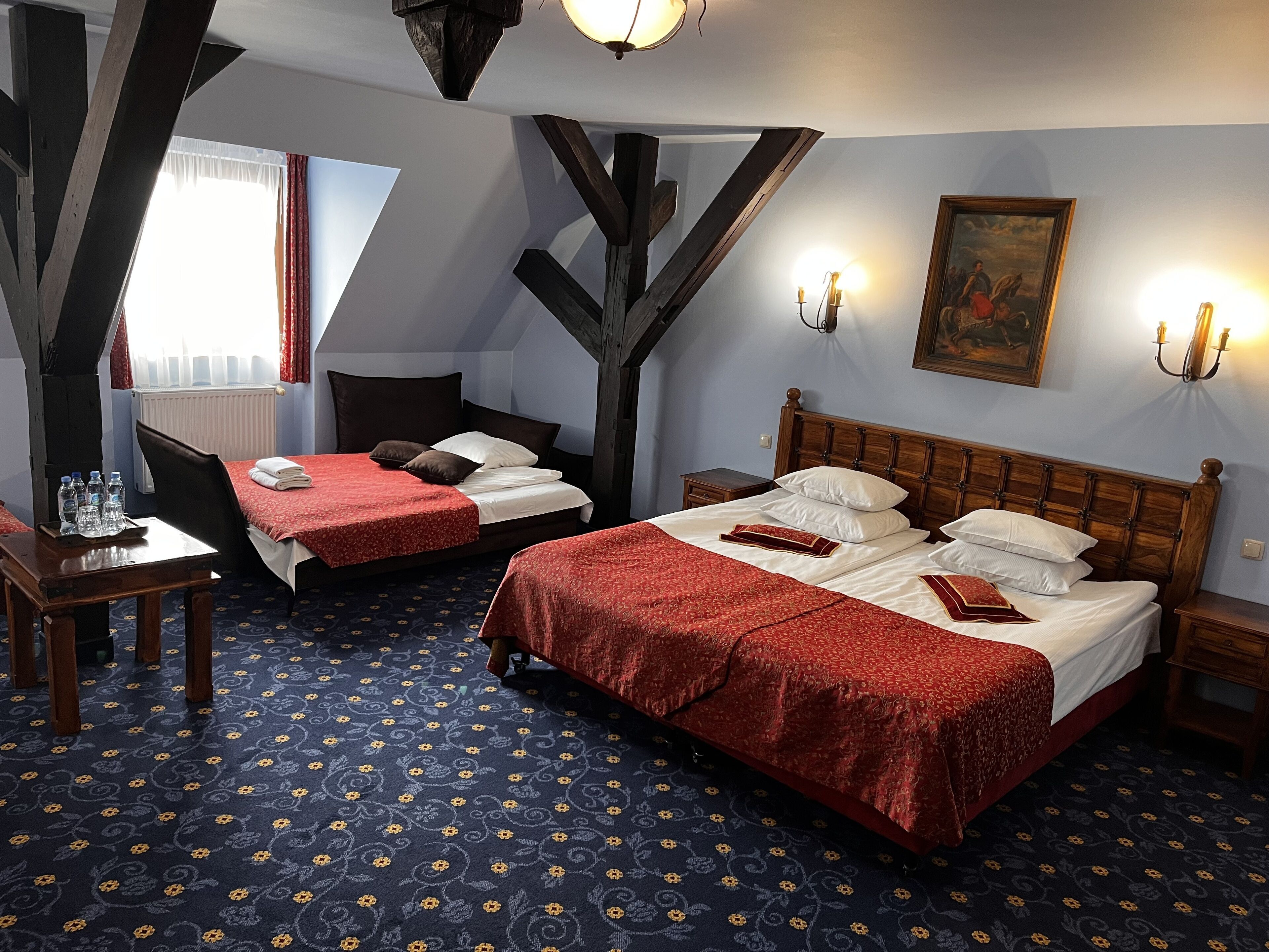 A hotel room with two beds, a wooden ceiling, a painting on the wall, and a window with curtains.