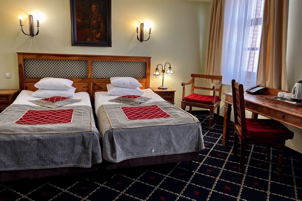 A hotel room with two single beds, a wooden desk with a chair, a telephone, and a lamp.