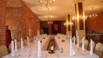 A banquet hall with long tables set for a formal event, featuring white tablecloths, candles, and gold chairs.