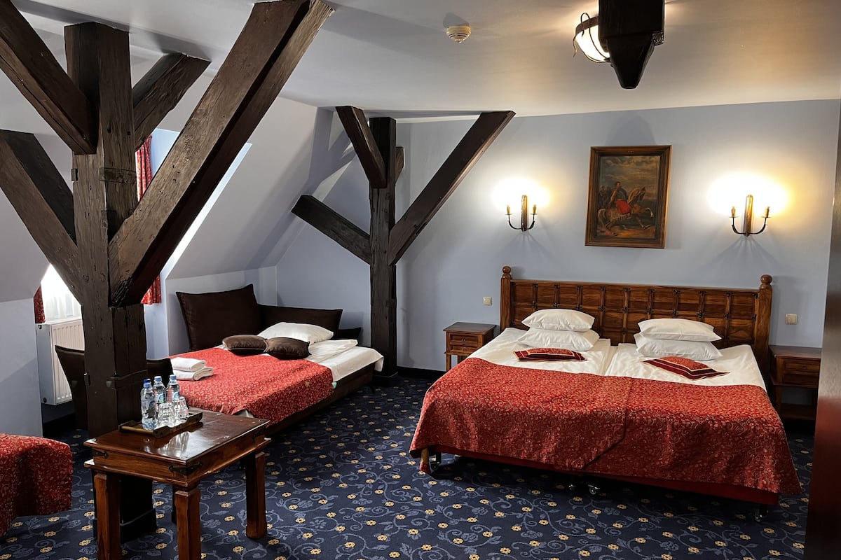 A room with two beds, a wooden ceiling, a patterned carpet, and a painting on the wall.