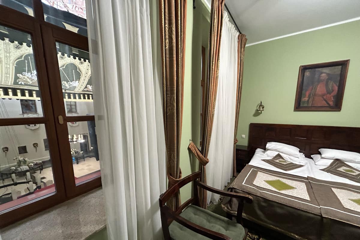 A hotel room with a bed, a chair, a window with curtains, and a painting on the wall.