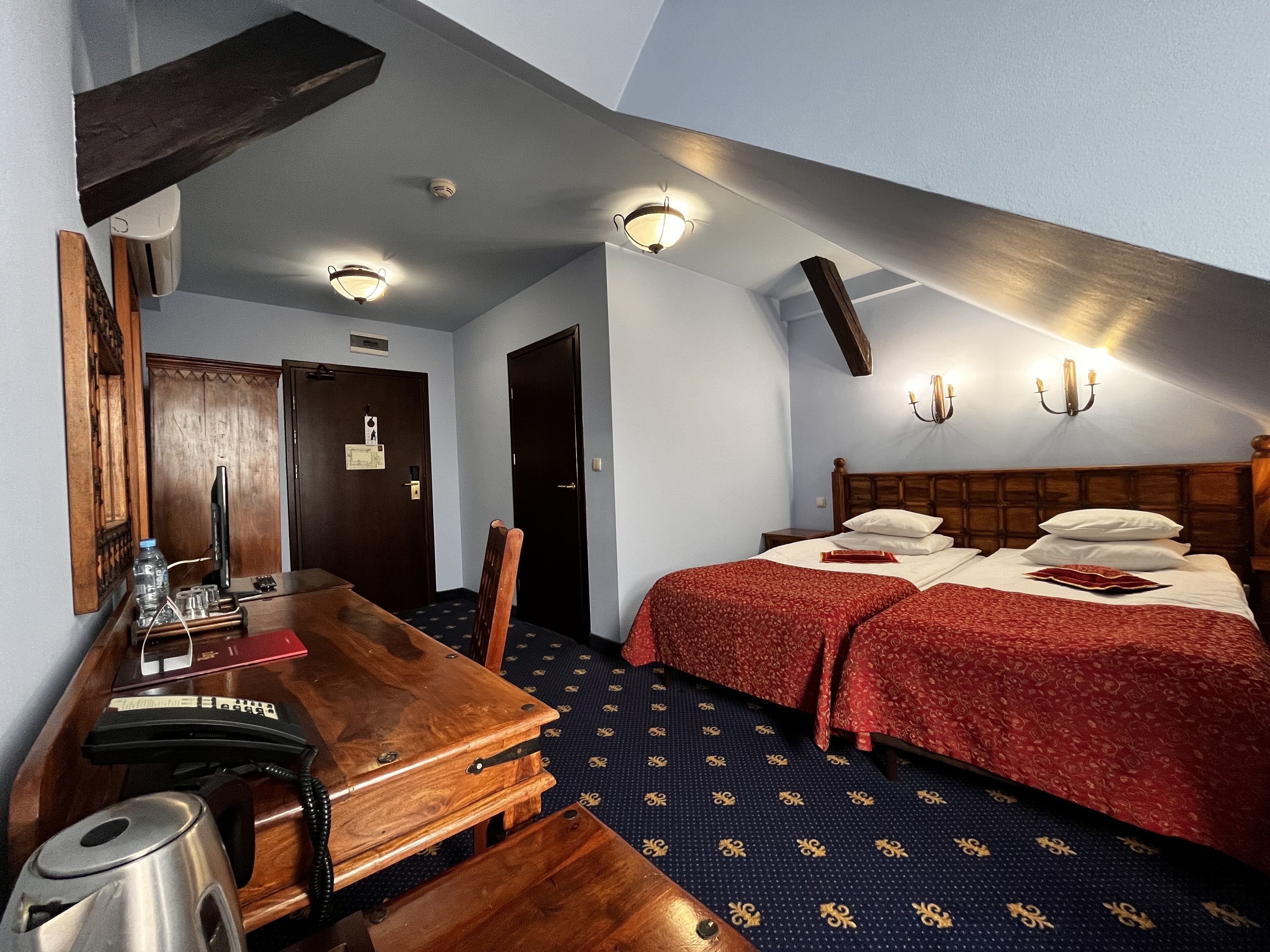 A hotel room with a wooden desk, two beds with red bedding, a patterned carpet, and a sloped ceiling with wall-mounted lights.