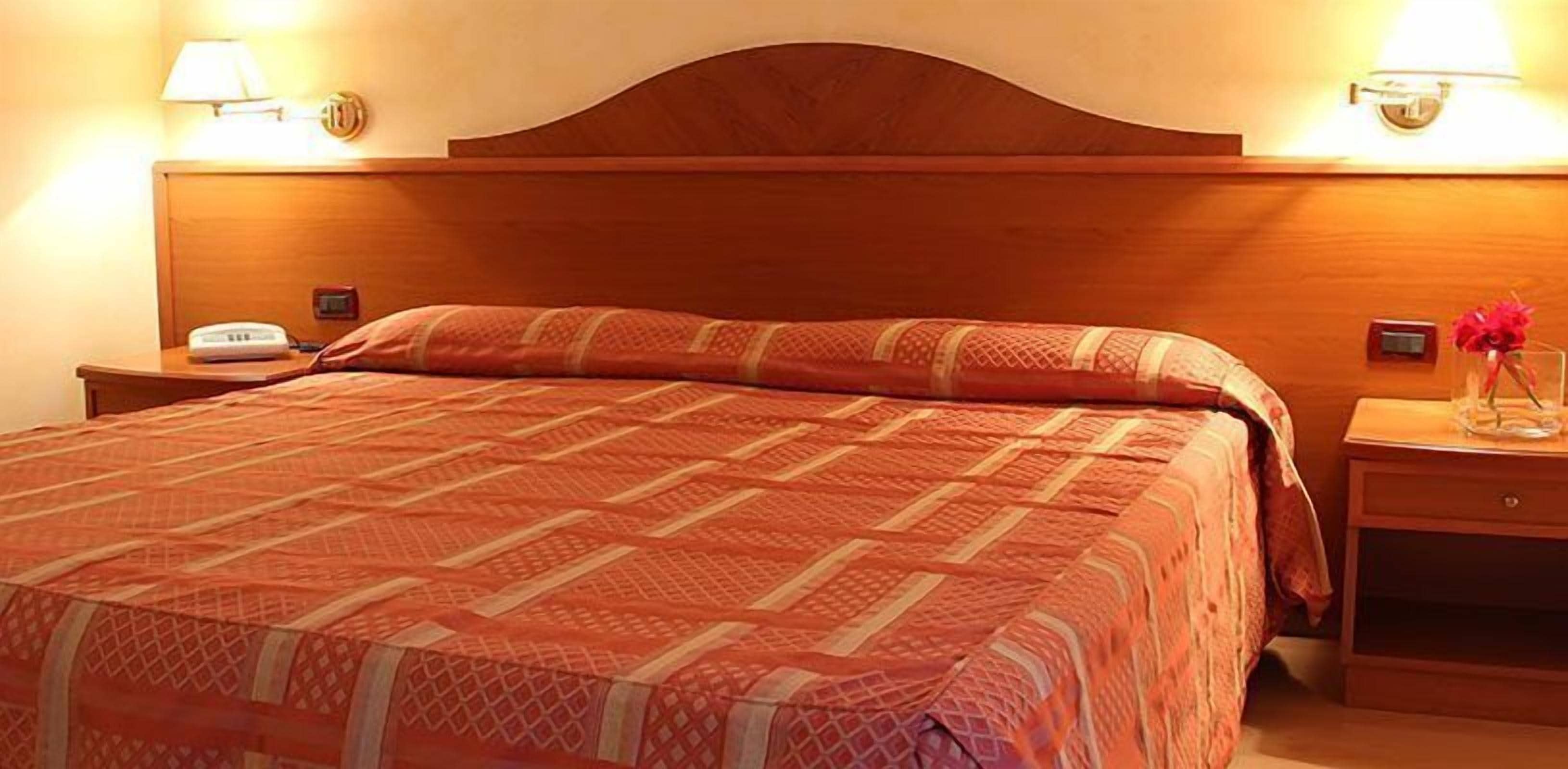 A hotel room with a wooden headboard, a patterned bedspread, a bedside table with a lamp, and a vase with flowers.