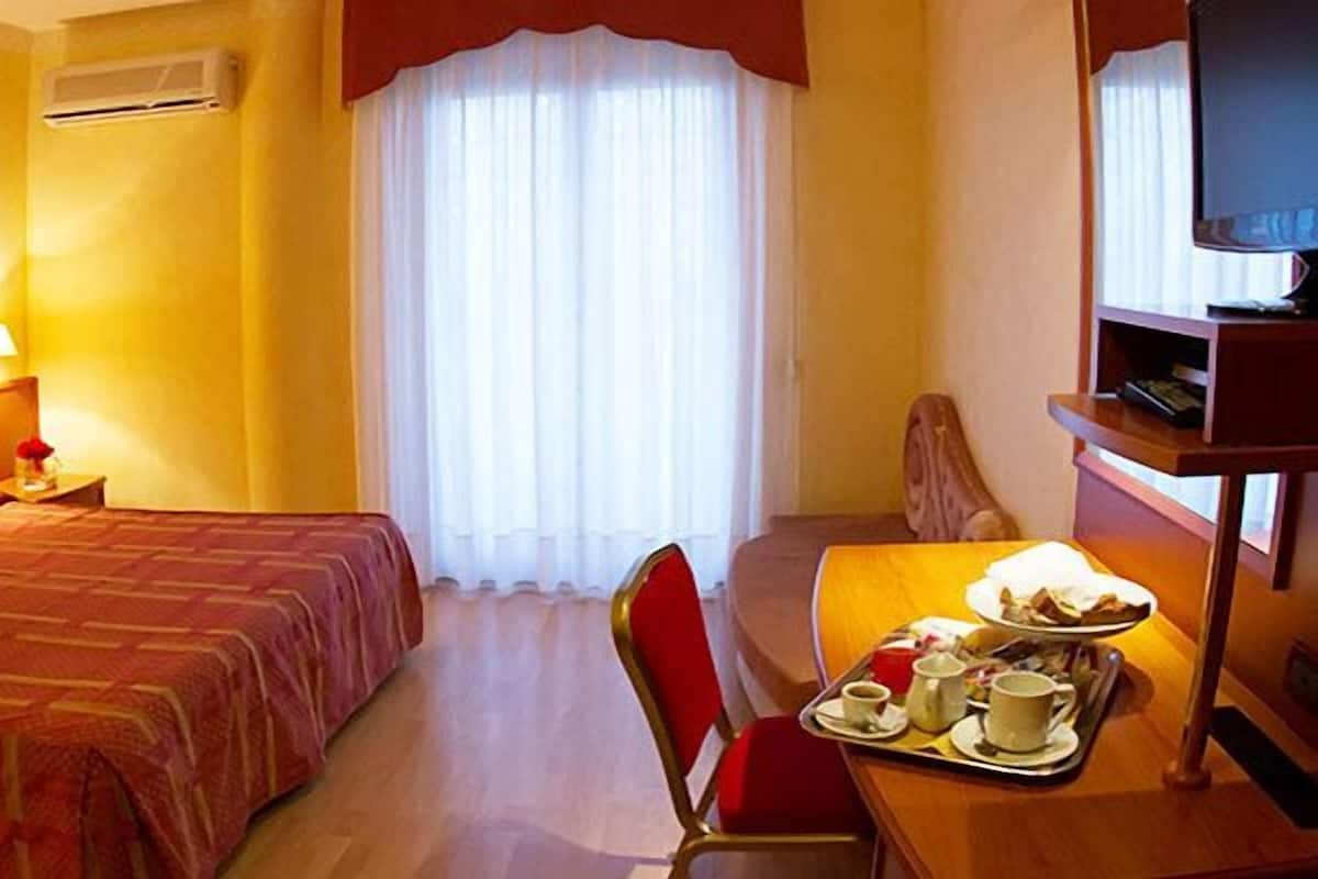 Une chambre d’hôtel avec un lit, un bureau sur lequel se trouve un plateau avec des articles pour le petit-déjeuner, une chaise, un téléviseur et une fenêtre avec des rideaux.