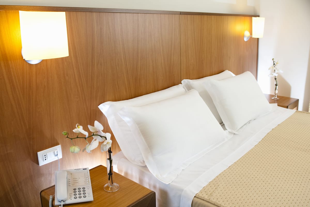 Una habitación de hotel con cabecera de madera, cama con sábanas blancas, mesita de noche con un teléfono y un jarrón de flores.