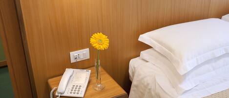 Habitación de hotel con una mesita de noche de madera, una cama blanca y una flor amarilla en un florero de cristal.