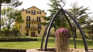 A yellow building with a balcony, surrounded by greenery and a stone fountain with a pink flower. - Hotel Residence Sant'Uberto (Roccastrada)