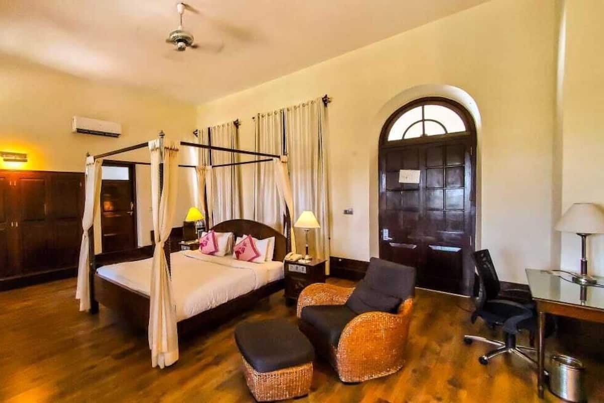 Uma cama com dossel e cortina, uma escrivaninha com cadeira, e um pequeno espaço de estar com poltrona e banquinho.