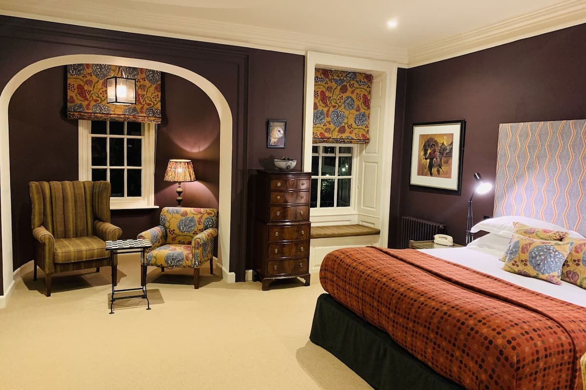 A bedroom with a bed, a chair, a dresser, a lamp, and a window with a patterned curtain.