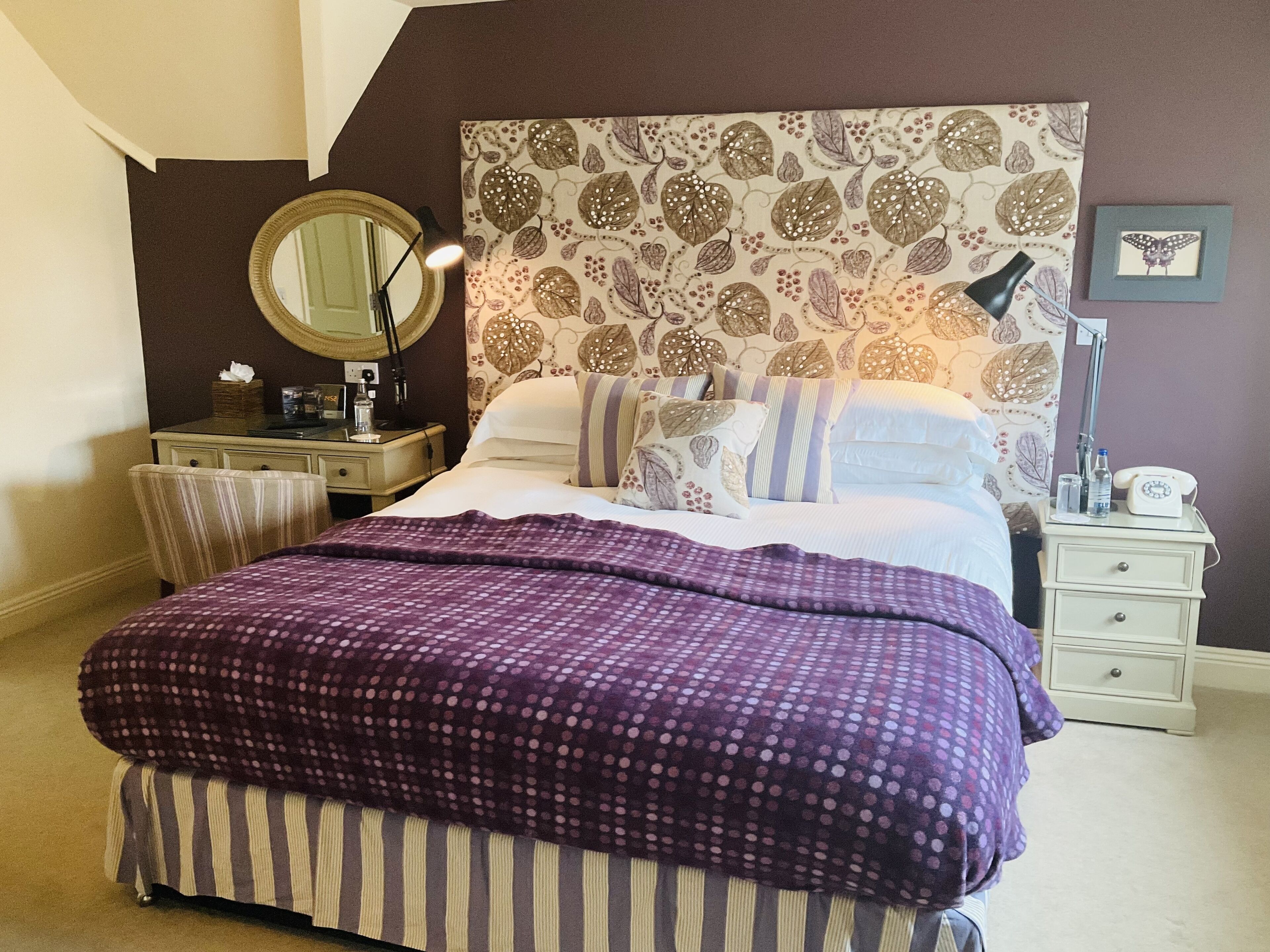 Quarto com cama, mesa de cabeceira, cômoda, espelho e parede com padrão de folhas.
