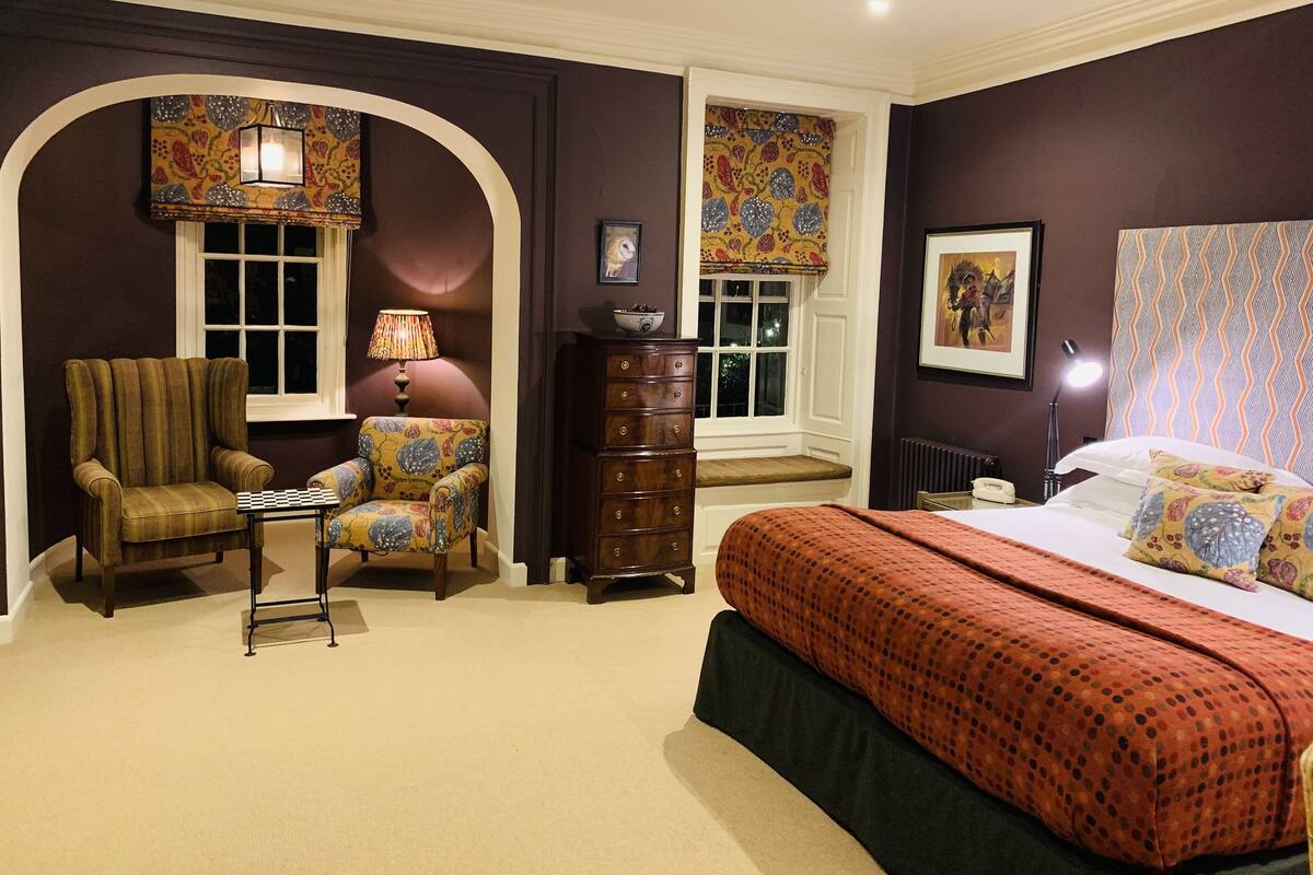 A bedroom with a large bed, a chair, a dresser, and a window with patterned curtains.