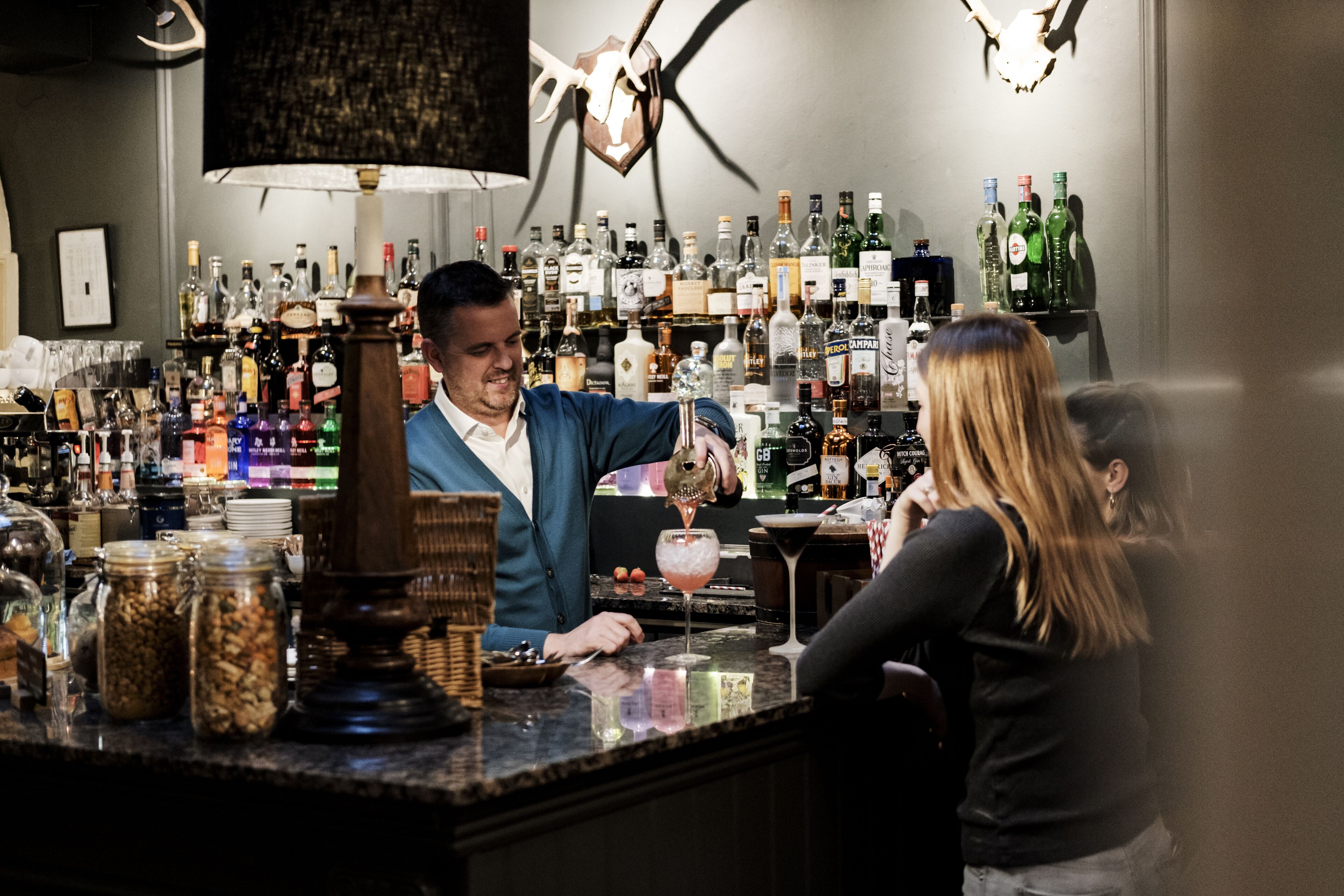 A bar area with a bartender preparing a drink, shelves stocked with various bottles, and a counter with jars.