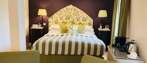 A hotel room with a large bed, two bedside lamps, a desk with a coffee maker, and two green armchairs.