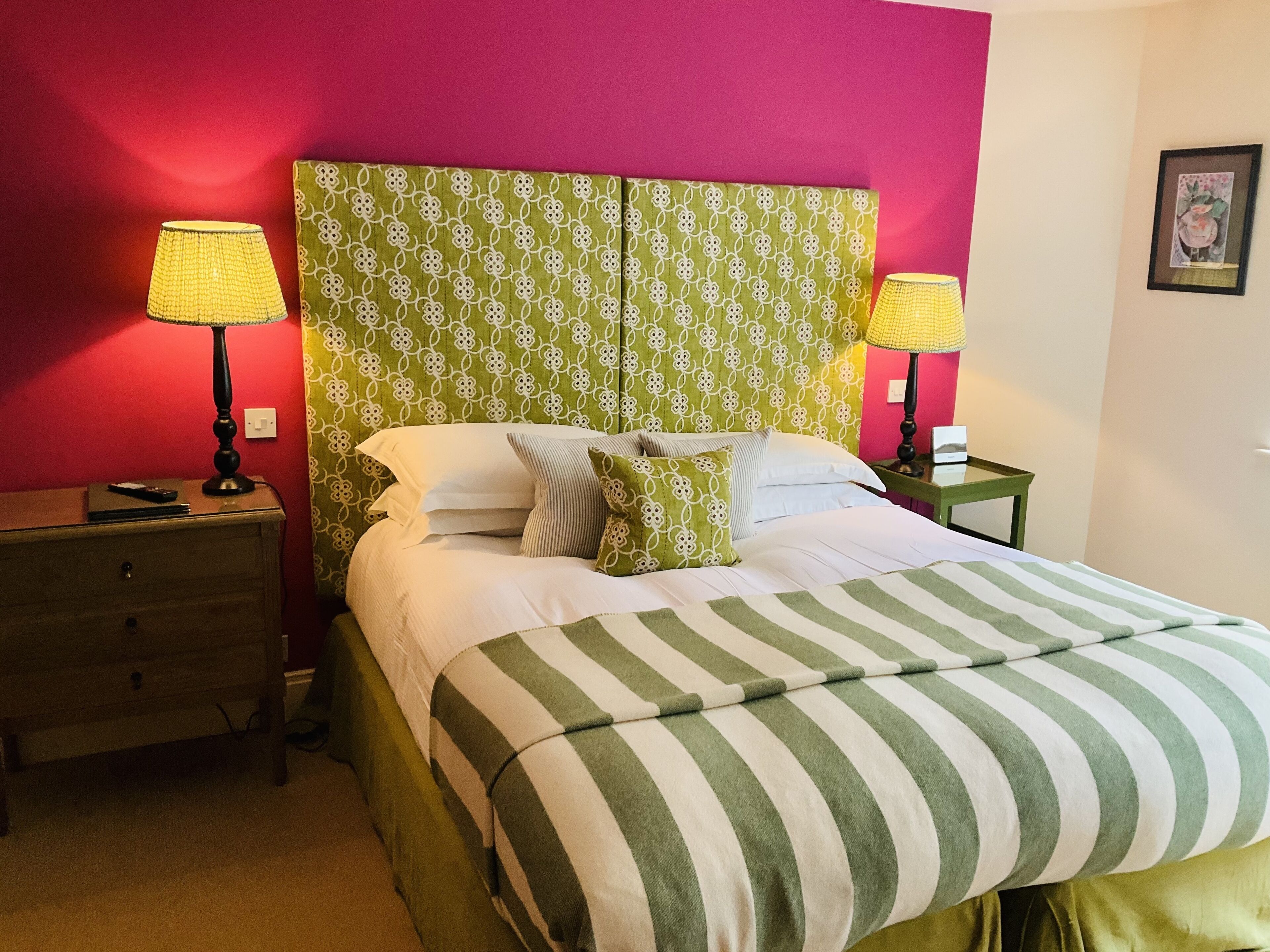 Cama bem arrumada com roupa de cama listrada, duas luminárias de cabeceira, cabeceira estampada em verde e uma parede de destaque vermelha.