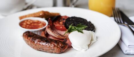 A plate of breakfast food including sausages, eggs, tomatoes, and toast, with a glass of orange juice in the background.