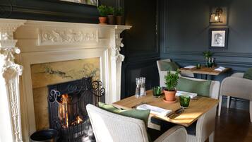 A dining area with a fireplace, wicker chairs, a wooden table, and potted plants.